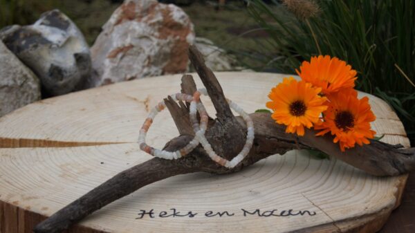 twee veelkleurige maansteen armbanden met chips kralen, hangend aan drijfhout op een houten schijf met logo 'Heks en Maan', naast oranje bloemen en stenen