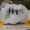 drie donkere lavasteen hangers aan zilverkleurige oogjes, hangend in een witte kwartskristal geode, omringd door oranje bloemen op een houten schijf met logo 'Heks en Maan