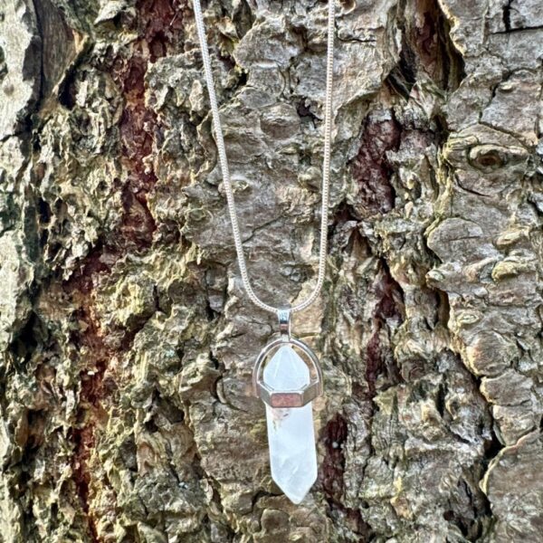 puntvormige bergkristal hanger met zilverkleurige ketting hangend tegen een ruwe boomstam