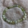 Een armband gemaakt van ronde, gepolijste olijf jade kralen in verschillende groen- en wittinten, liggend op een zachte, crèmekleurige ondergrond.
