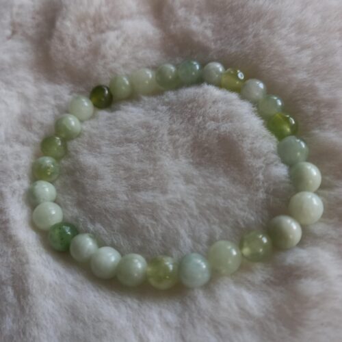 Een armband gemaakt van ronde, gepolijste olijf jade kralen in verschillende groen- en wittinten, liggend op een zachte, crèmekleurige ondergrond.
