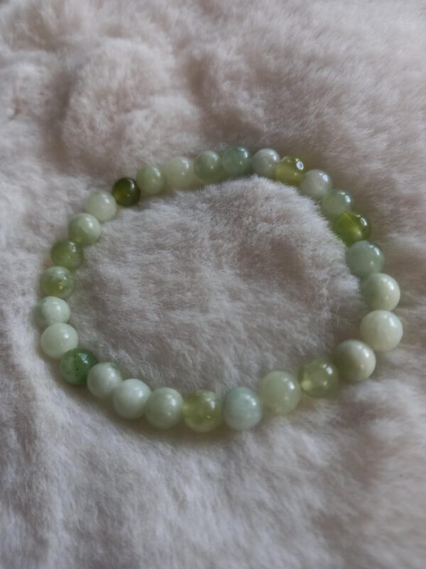 Een armband gemaakt van ronde, gepolijste olijf jade kralen in verschillende groen- en wittinten, liggend op een zachte, crèmekleurige ondergrond.