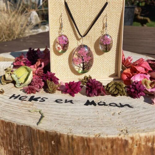Een felroze Tree of Life sieradenset (ketting en oorbellen) met hars en gedroogde bloemen, gepresenteerd in een doosje op een boomstronk met bloemblaadjes en de tekst 'Heks en Maan'.