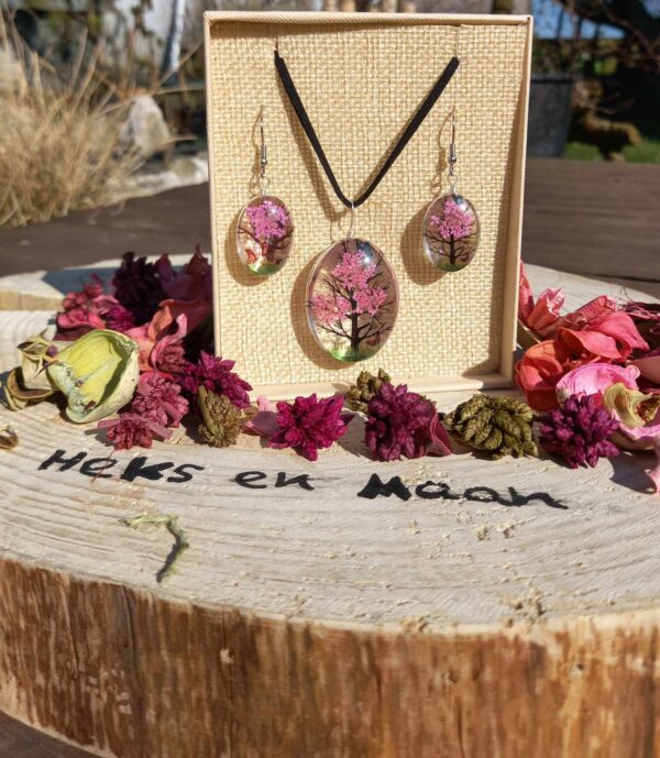 Een felroze Tree of Life sieradenset (ketting en oorbellen) met hars en gedroogde bloemen, gepresenteerd in een doosje op een boomstronk met bloemblaadjes en de tekst 'Heks en Maan'.